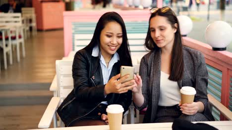 Hübsche-junge-Damen,-die-Freunde-benutzen-moderne-Smartphone-sitzen-am-Tisch-im-Café-mit-Kaffee-zum-Mitnehmen.-Glückliche-Mädchen-sind-Bildschirm-berühren,-Fotos-anschauen-und-lachen.