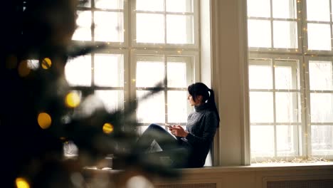 Glückliche-junge-Frau-nutzt-Smartphone-sitzen-auf-Fensterbank-am-Weihnachtstag-zu-Hause-entspannen-und-genießen-modernen-Technologie.-Menschen-und-Geräte-Konzept.