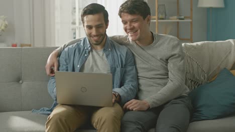 Entzückende-männliche-Gay-paar-verbringen-Zeit-zu-Hause.-Sie-sitzen-auf-einem-Sofa-und-das-Notebook-nutzen.-Sie-ansehen-lustige-Online-Videos.-Partner-legt-seine-Hand-um-seine-Geliebte.-Zimmer-ist-Modern-eingerichtet.