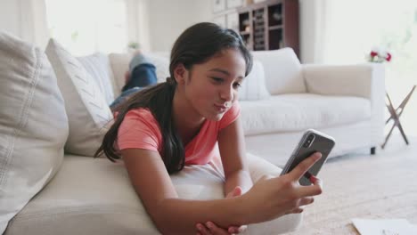 Happy-teenage-Hispanic-girl-lying-on-a-sofa-at-home-using-smartphone,-low-angle,-close-up