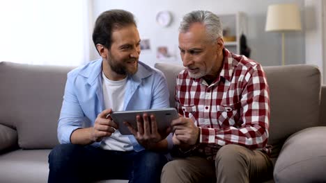 Retiree-father-and-middle-aged-son-scrolling-tab,-looking-and-smiling-to-camera