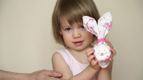 Portrait-of-little-pretty-smiling-caucasian-girl-holds-chicken-egg-decorated-for-Easter-bunny,-with-painted-muzzle-and-ears.