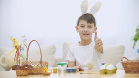 Smiling-boy-is-colorizing-an-Easter-egg-with-a-help-of-paint-brush.,-wearing-bunny-ears,-then-raises-his-left-thumb-finger-up.