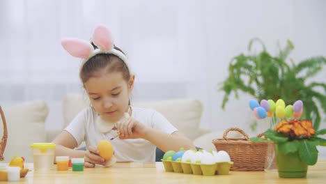 Intersted-and-concentrated-girl-is-finishing-to-colourize-an-Easter-egg-in-an-yellow-colour,-sitting-at-the-table.