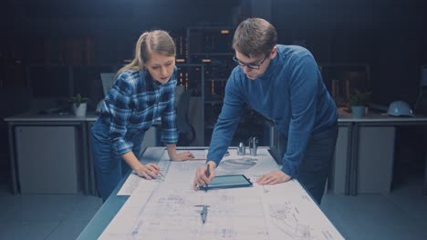 En-la-instalación-de-ingeniería-de-diseño-industrial-oscuro-hombres-y-mujeres-ingenieros-hablan-y-trabajan-en-un-plano-utilizando-la-mesa-de-conferencias.-En-los-planos-de-escritorio,-borradores-y-componentes-de-motor-eléctrico,-piezas