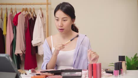 Beauty-Blogger-präsentieren-Beauty-Kosmetik-sitzen-vor-dem-Tablet-für-die-Aufnahme-von-Video.-Glücklich-schöne-junge-asiatische-Frau-verwenden-Kosmetik-Rezension-Make-up-Tutorial-übertragen-Live-Video-an-soziale-Netzwerk.