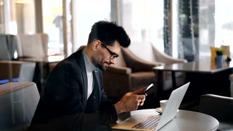 Smiling-business-man-touching-smartphone-screen-enjoying-social-media-in-cafe