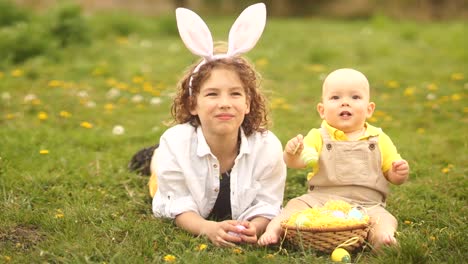 Two-brothers,-a-teenager-and-a-baby-are-resting-in-a-park-on-a-glade.-Easter-concept,-bunny-ears,-easter-basket-and-eggs.-A-dog-runs-past-them