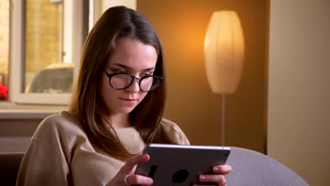 Retrato-en-primer-plano-de-joven-atractiva-mujer-caucásica-en-gafas-usando-la-tableta-sentada-en-el-sofá-en-el-interior-de-un-acogedor-apartamento