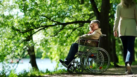 Tilt-down-of-pensive-mature-man-with-beard-sitting-in-wheelchair-near-lake-in-park-alone-and-looking-at-view.-Unrecognizable-woman-passing-by-him-on-windy-day