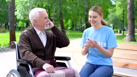 Happy-young-woman-giving-earphone-old-disabled-man,-supporting-hospital-patient