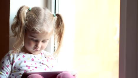 Blonde-little-girl-using-tablet-pc.-Sitting-on-the-window-sill.-Child-smiling-and-looking-at-camera.-Child-plays-on-the-PC