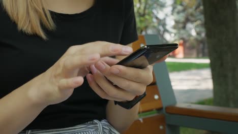 Close-up-shot-of-a-young-female-scrolling-and-typing-on-his-black-phone-outside-in-city-park-in-summer.-Woman-texting-and-chatting-message-on-smartphone-outdoors.