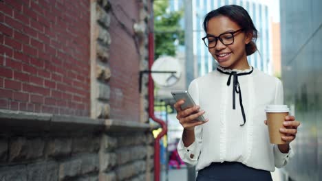 Smiling-African-American-woman-using-smartphone-holding-coffee-walking-outdoors