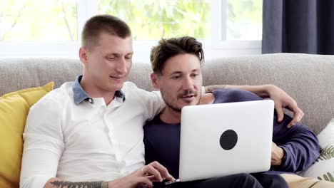 Pareja-gay-relajándose-en-el-sofá-usando-la-computadora-portátil.-Prueba-a-comprar-en-línea.