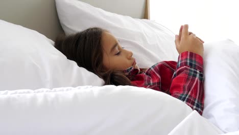 Nettes-Mädchen-liegt-auf-dem-Bett-und-schaut-auf-Smartphone-und-Social-Media-Unterhaltung.-Menschen-Lebensstil-und-Bildung.-Kinder--und-Kinderkonzept.-Mädchen-wischen-Touchscreen-auf-Handy.-Technologie-Thema