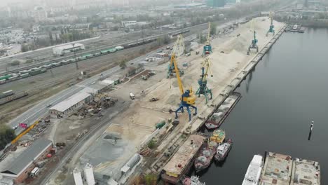 Aerial-view-of-port-working-cranes-extracting-sand-from-iron-barge-and-scow-on-river-bank-in-industrial-dirty-city-in-smog