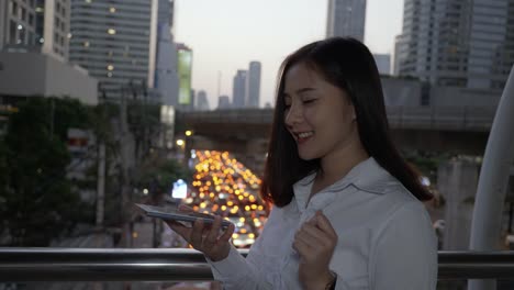 Young-smiling-business-asian-woman-using-smart-phone-in-the-modern-city-center-in-the-twilight-of-Bangkok-Thailand.-She-uses-her-finger-to-touch-the-screen.-For-wearing-holograms-You-design