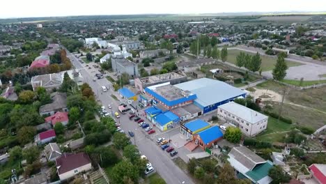 Exciting-bird`s-eye-view-of-a-wide-highway-with-moving-cars-going-through-a-green-city-with-large-warehouses,-small-buildings-and-parks-in-summer