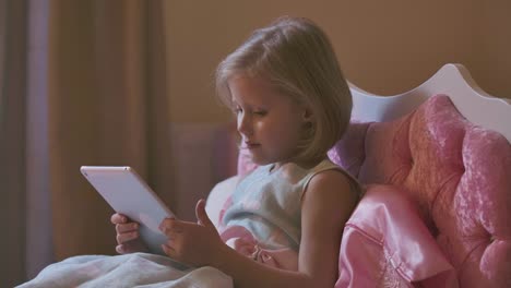 Portrait-of-pretty-blond-Caucasian-girl-lying-in-pink-bed-and-using-tablet.-Modern-child-watching-cartoons-before-going-to-bed.-Resting-at-home,-carefree-childhood.