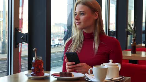 Eine-junge-attraktive-Frau-sitzt-an-einem-Tisch-mit-Essen-in-einem-Café-und-arbeitet-auf-einem-Smartphone-mit-einem-Lächeln,-schließlich-schaut-aus-dem-Fenster