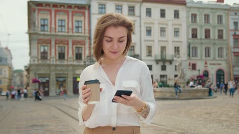 The-girl-is-texting-on-the-phone.-She-is-standing-and-smiling.-She's-holding-a-cup-of-coffee.-The-sun-is-shining-and-people-are-walking-in-the-background.-4K