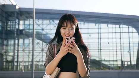 Close-view-of-jocund-happy-25-aged-asian-lady-with-long-hair-and-stylish-clothes-which-using-her-smartphone-near-modern-airport-building