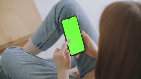 Standpunkt-der-Frau-im-modernen-Zimmer-sitzen-auf-einem-Stuhl-mit-Telefon-mit-grünen-Mock-up-Bildschirm-Chroma-Key-Surfen-Internet-beobachten-Inhalte-Videos-Blogs-Tippen-auf-Center-Screen