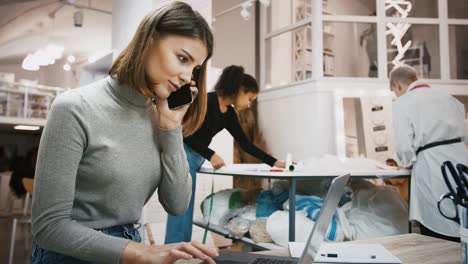 Woman-accepting-order-from-client-by-mobile-phone,-typing-on-laptop-working-at-sewing-workshop,-mixed-race-tailors-working-nearby