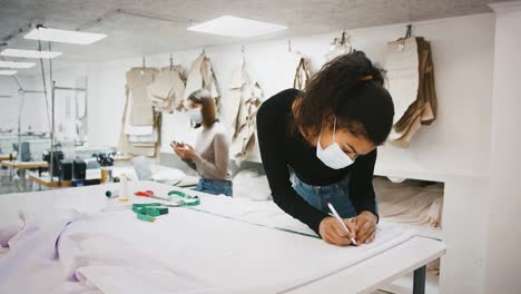 Dark-skinned-female,-tailor-of-sewing-workshop-in-protective-mask-taking-measurements-of-fabric-by-tape-measure,-coronavirus