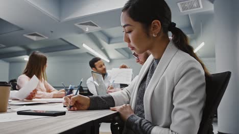 Mujer-asiática-escribiendo-información-escuchada-de-jefe-que-explica-detalles-del-proyecto-de-startup-a-diversos-colegas,-sentados-en-la-oficina-moderna