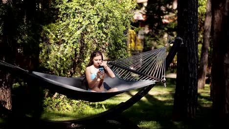 A-girl-in-a-blue-swimsuit-uses-her-phone-and-swings-in-a-hammock-in-nature.