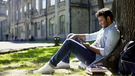 Multiracial-young-man-sitting-under-tree,-scrolling-mobile-phone-screen,-smiling