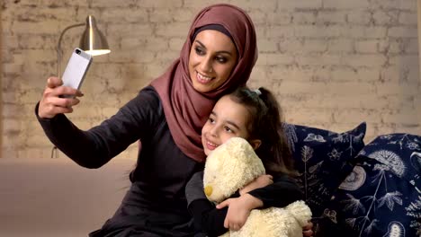 Junge-schöne-Mutter-Hijab-mit-kleinen-Mädchen-auf-der-Couch,-lächelnd,-Smartphone-nutzt,-macht-Selfie,-Kuscheln,-kleines-Mädchen-mit-Teddybär,-home-Komfort-im-Hintergrund-50-fps