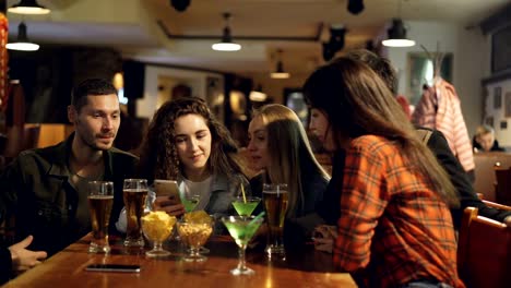 Joven-mujer-muestra-algo-gracioso-en-la-pantalla-de-su-smartphone-a-sus-amigos-cuando-están-sentados-en-la-mesa-de-café.-Sus-compañeros-riendo-y-hablando-de-ello.