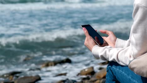 Mujer-está-leyendo-algo-en-el-teléfono-sentado-en-la-orilla-del-mar-en-otoño.-Manos-cerca.