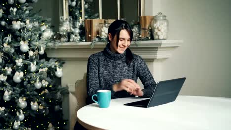 Glückliches-Mädchen-ist-mit-Laptop-zu-Weihnachten-Tag-sitzen-am-Tisch-in-der-Nähe-von-Neujahr-Baum-geschmückt-und-tippen-auf-Bildschirm-senden-von-E-mails-an-Freunde.