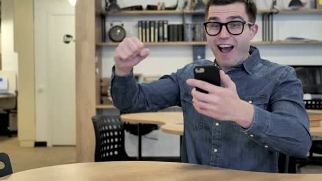 Creative-Young-Man-in-Glasses-Excited-for-Success-while-Using-Smartphone