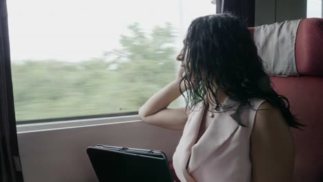 Daydreaming-young-corporate-woman-looking-out-window-on-train-and-texting-on-tablet-pc