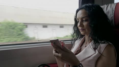 Joven-pasajera-viajera-viajando-en-tren-escuchando-música-usando-auriculares-y-chateando-en-un-smartphone-conectado-por-Internet-inalámbrico