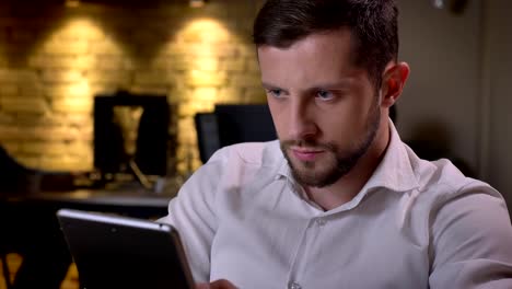 Closeup-portrait-of-young-focused-caucasian-businessman-browsing-on-the-tablet-in-the-office-on-workplace