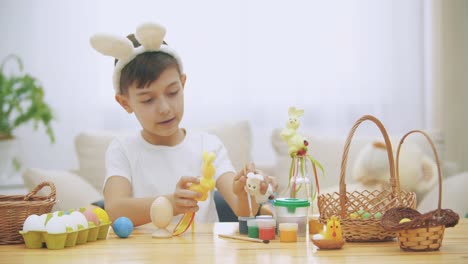 Young-adorable-boy-is-sitting-at-the-table-full-of-Easter-decorations-and-is-playing-with-Easter-bunnies-in-his-hands.-Bunnies'-discussion.-Who-would-paint-an-egg-Bunny-chooses-green-colour.-Bunny-theatre.