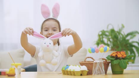 Pequeña-chica-linda-se-está-divirtiendo-pintando-con-conejito-de-Pascua,-que-tiene-las-mismas-orejas,-como-ella.-La-chica-sonriente-está-expandiendo-las-orejas-del-conejito-y-luego-lo-abraza.-Chica-con-un-punto-de-belleza-en-la-cara-y-está-sonriendo-am