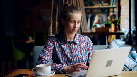 Happy-young-lady-working-with-laptop-in-cafe-typing-sitting-at-table-alone