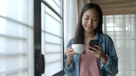 Attractive-portrait-smiling-young-asian-woman-drink-coffee-and-typing-mobile-phone-and-scrolls-through-social-media-feed-in-smartphone-standing-beside-window-at-home-office.