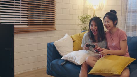 Lesbian-lgbt-women-couple-play-games-at-home,-Asian-female-using-joystick-having-funny-happy-moment-together-on-sofa-in-living-room-in-night.-Young-lover-football-fan,-celebrate-holiday-concept.