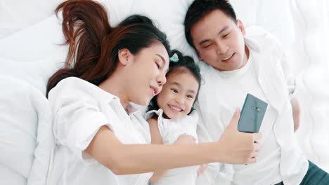 Asiatische-Familie-lacht-und-schaut-in-Smartphone,-während-auf-dem-Bett-im-Schlafzimmer-liegen