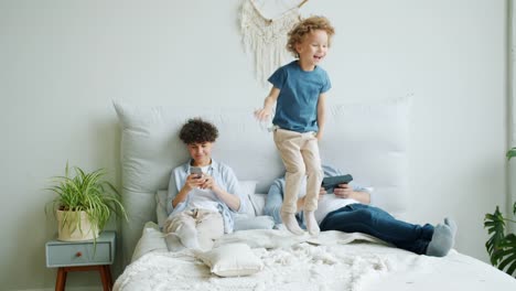 Joyful-kid-jumping-on-bed-while-mother-using-smartphone-father-holding-tablet