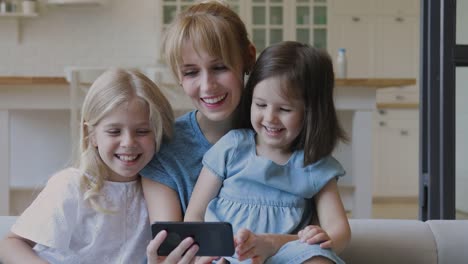 Madre-feliz-y-las-hijas-pequeñas-riendo-viendo-dibujos-animados-en-el-teléfono-inteligente