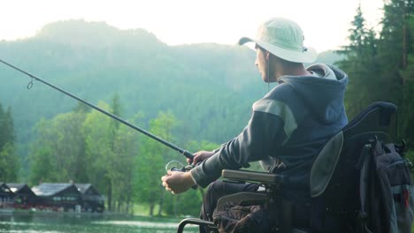 Resolución-4k-de-pescador-discapacitado-en-una-silla-de-ruedas-eléctrica-pescando-en-un-hermoso-lago-cerca-del-bosque-y-la-montaña-en-la-parte-posterior,-en-la-puesta-de-sol,-verano